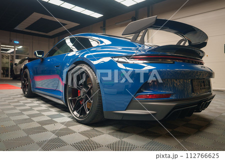 A gleaming blue Porsche 911 GT3 with a prominent rear wing and Porsche lettering is showcased indoors, reflecting lights on its polished surface and sporty alloy wheels with red calipers. 112766625