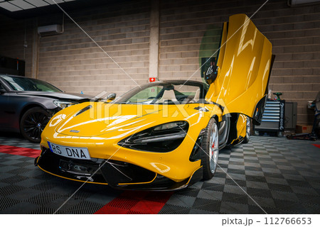 A vibrant yellow McLaren 765LT with upwardopening dihedral doors is displayed in a tuning garage, featuring black aerodynamic accents and multispoke wheels against a checkered floor. 112766653