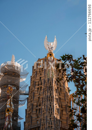 Closeup of Sagrada Familia's tower in Barcelona, adorned with Gaudi's signature motifs, a backlit cross, and dove sculptures against a clear sky, framed by foliage. 112766698