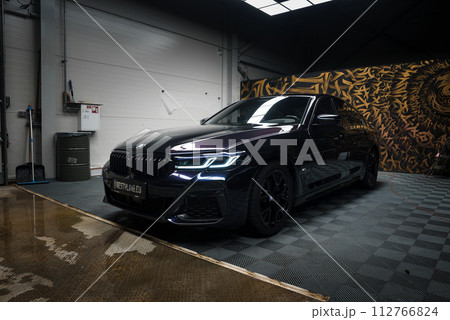 A sleek black BMW with headlights on is centered in a tuning garage with a checkered floor, wet patches reflecting light, and walls featuring edgy graffiti, indicating a highperformance service area. A sleek black BMW with headlights on is centered in a tuning garage with a checkered floor, wet patches reflecting light, and walls featuring edgy graffiti, indicating a highperformance service area. 112766824