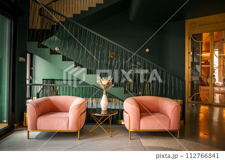 Elegant spa resort interior in Druskininkai, Lithuania, featuring deep green walls, plush pink armchairs with gold accents, a modern staircase with black rods, and a welllit conference area. Elegant spa resort interior in Druskininkai, Lithuania, featuring deep green walls, plush pink armchairs with gold accents, a modern staircase with black rods, and a welllit conference area. 112766841