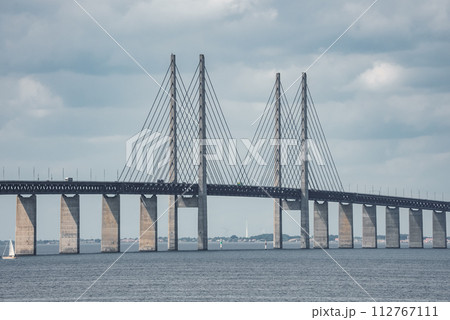 Capturing the grandeur of the Oresund Bridge, this image features its towering pylons and harplike cables over a navigable waterway, with a sailboat and overcast skies. 112767111