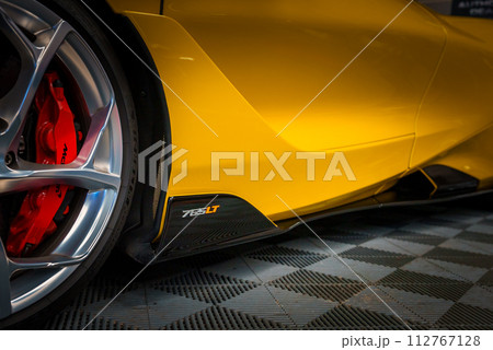 Closeup of a yellow McLaren 765LT's sleek side profile near the rear wheel, highlighting its aerodynamic design, red brake caliper, and silver alloy wheel on a checkered floor. 112767128
