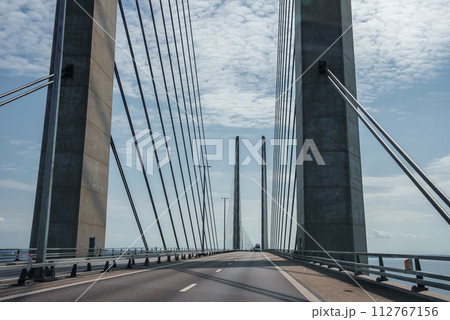 Capturing the sleek engineering of the Oresund Bridge, this image features its towering concrete supports and harplike cables under a clear blue sky, on a trafficfree multilane highway. Capturing the sleek engineering of the Oresund Bridge, this image features its towering concrete supports and harplike cables under a clear blue sky, on a trafficfree multilane highway. 112767156