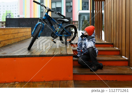 A Caucasian boy dressed in a winter jacket walks with a bicycle around the city 112771261