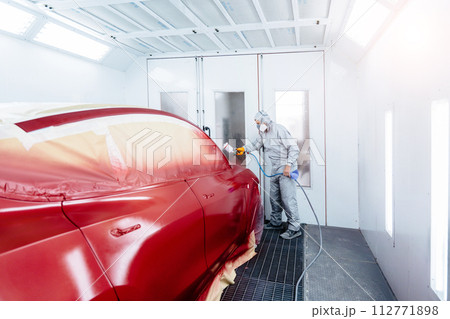 Wide angle of automobile repairman painter in protective workwear and respirator painting car body in paint chamber. Wide angle of automobile repairman painter in protective workwear and respirator painting car body in paint chamber. 112771898