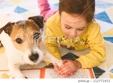 Small girl learning to read. Kid is reading a book to her pet dog. 112771915