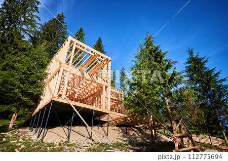 Residential building with wooden frame in process of being built near a forest. Beginning of new construction of comfortable cottage. Idea of ecological construction, contemporary architectural design Residential building with wooden frame in process of being built near a forest. Beginning of new construction of comfortable cottage. Idea of ecological construction, contemporary architectural design 112775694