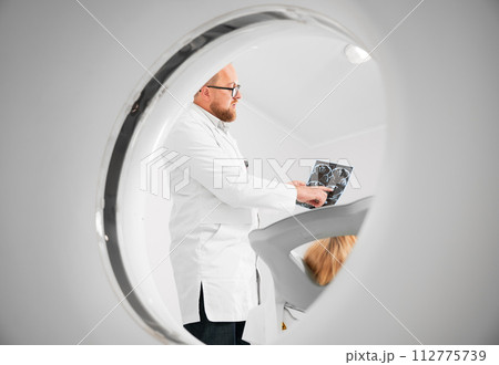 Medical computed tomography or MRI scanner. Doctor holding and explaining results of MRI. Woman patient lying on couchette. Concept of medicine, healthcare and modern diagnostics. 112775739