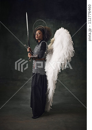 Young African woman, medieval warrior with angel wings, halo above head and chainmail holding sword against vintage dark background. 112776460