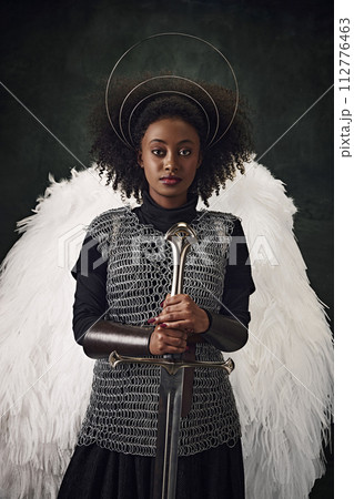 Young African woman with angel wings, halo above head and chainmail holding sword against vintage dark background. 112776463