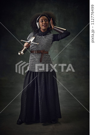 Young African woman, medieval warrior in chainmail armor with sword listening to music in headphones and singing against vintage green background. 112776499