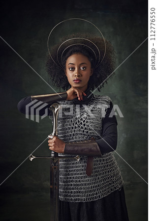 Brave young African woman, medieval warrior in chainmail armor with halo-like rings above head leaning sword on dark green vintage background Brave young African woman, medieval warrior in chainmail armor with halo-like rings above head leaning sword on dark green vintage background 112776550