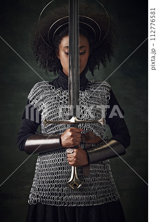 Portrait of young African woman, medieval warrior in chainmail armor with halo-like rings above head holding sword close to face on dark vintage background Portrait of young African woman, medieval warrior in chainmail armor with halo-like rings above head holding sword close to face on dark vintage background 112776581