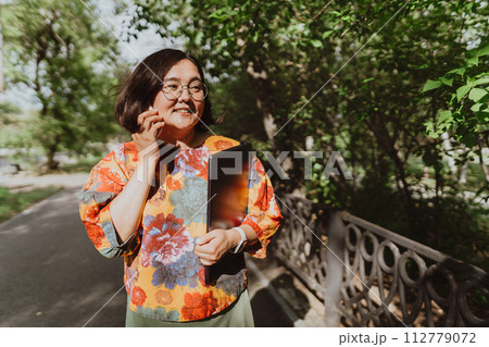 Happy woman chatting on the phone holding a laptop walking in the park Smiling freelancer wearing a colorful blouse using smartphone outdoors 112779072