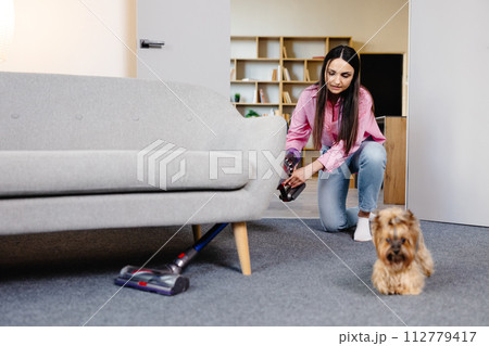 Young woman uses cordless vacuum cleaner to clean home carpet, and a dog is running nearby. Modern easy cleaning 112779417