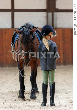 Horse riding school. Little children girls at group training equestrian lessons in indoor ranch horse riding hall. Cute little beginner blond girl kid in helmet sitting on brown horse horseback 112781243