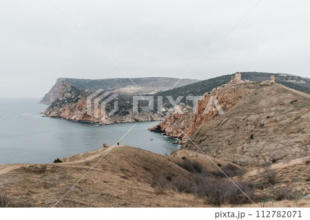 Woman stands on cliff overlooking ocean, cloudy day provides soft lighting. She's wearing a yellow sweater wearing dark pants. Landscape features rugged terrain, calm waters. Woman stands on cliff overlooking ocean, cloudy day provides soft lighting. She's wearing a yellow sweater wearing dark pants. Landscape features rugged terrain, calm waters. 112782071