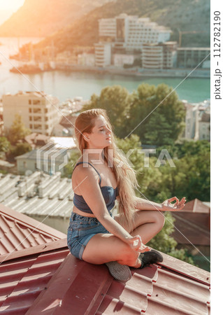 Woman sits on rooftop, enjoys town view and sea mountains. Peaceful rooftop relaxation. Below her, there is a town with several boats visible in the water. Rooftop vantage point. 112782190