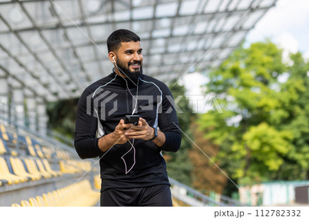 A smiling, bearded man wearing sportswear and earphones holds a smartphone while standing at an outdoor stadium. 112782332