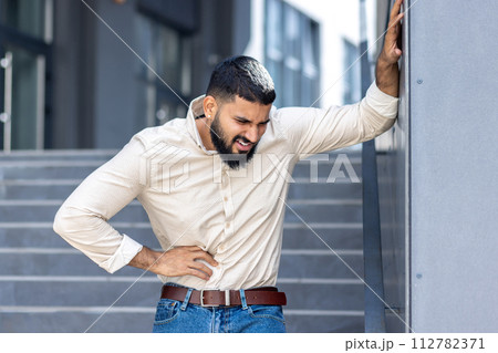 A young Muslim man in a shirt is standing outside an office building, leaning his hand on the wall and holding the side of his body, feeling severe pain in his stomach, grimacing from convulsions. 112782371