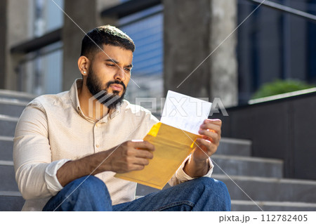 A serious young Indian man is sitting on the steps outside the building, holding an envelope and reading the letter and the results in a sad way. Close-up photo. 112782405