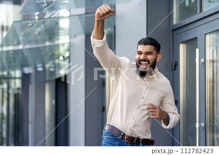 Indian young man standing near an office building and rejoicing in success raising his hand up and shouting with happiness, he got a job, a promotion, a business deal. Indian young man standing near an office building and rejoicing in success raising his hand up and shouting with happiness, he got a job, a promotion, a business deal. 112782423
