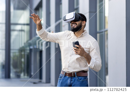 Photo of a young Muslim man in a shirt standing outside an office building wearing a virtual mask, holding a phone, talking online and gesturing with his hands. Photo of a young Muslim man in a shirt standing outside an office building wearing a virtual mask, holding a phone, talking online and gesturing with his hands. 112782425