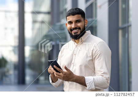 Portrait of a young Indian man in a casual shirt standing near an office center, holding a mobile phone and looking at the camera. Portrait of a young Indian man in a casual shirt standing near an office center, holding a mobile phone and looking at the camera. 112782457