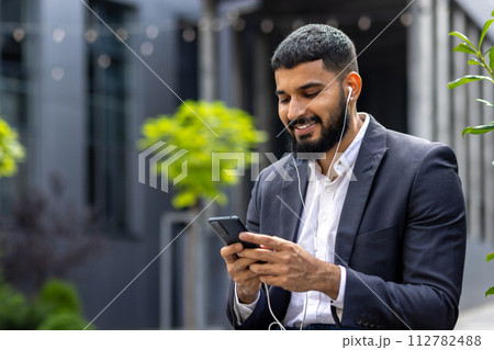 A modern businessman in a suit and earphones is engaged with his smartphone, smiling as he reads the screen, amidst an urban backdrop with verdant greenery. A modern businessman in a suit and earphones is engaged with his smartphone, smiling as he reads the screen, amidst an urban backdrop with verdant greenery. 112782488