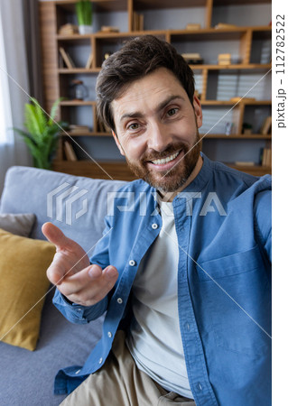 A relaxed man in casual attire sitting on a sofa, gesturing as if in the middle of a conversation, with a bookshelf in the background. 112782522