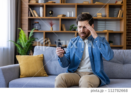 Stressed male sitting on a gray couch at home, expressing anxiety while looking at his mobile phone, with a hand on his forehead. 112782668