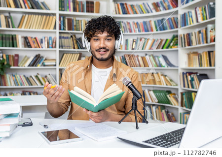 Confident blogger in white headset recording audio content while sitting with daybook by desk with professional micro. Islamic lecturer reading information for remote listeners in cyberspace. 112782746