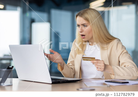 Shocked businesswoman holding a credit card looking at her laptop screen with confusion and surprise in an office setting. 112782979