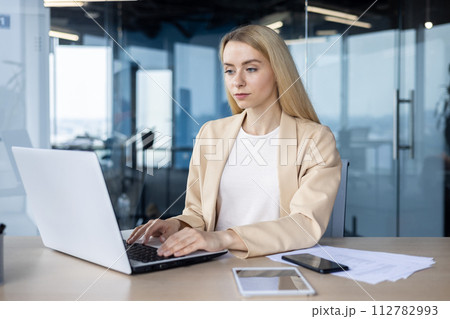 Professional woman in a beige blazer typing intently on a laptop at her workspace with smartphone and tablet in reach. Professional woman in a beige blazer typing intently on a laptop at her workspace with smartphone and tablet in reach. 112782993