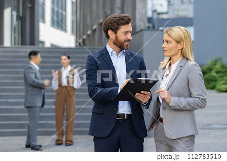 Two corporate professionals in suits engaged in a discussion with a clipboard outside a modern office building. Two corporate professionals in suits engaged in a discussion with a clipboard outside a modern office building. 112783510