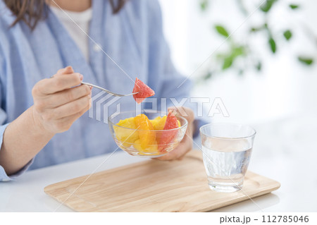 水のグラスを手にフルーツを食べるミドル女性の手元 112785046