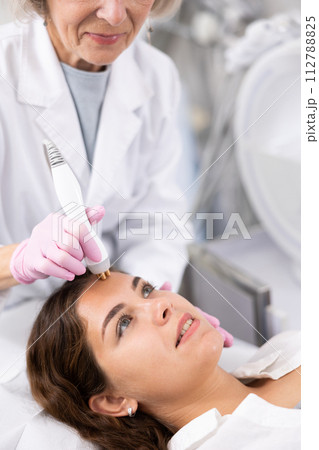 Elderly beautician doctor doing RF lifting procedure for flawless woman face laying in beauty salon. Hardware cosmetology. Elderly beautician doctor doing RF lifting procedure for flawless woman face laying in beauty salon. Hardware cosmetology. 112788825