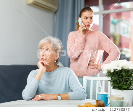 Upset senior woman sitting at table while irritated adult daughter talking on phone Upset senior woman sitting at table while irritated adult daughter talking on phone 112788877
