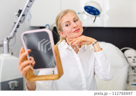 Female client looking in mirror satisfied with results of cosmetology procedure Female client looking in mirror satisfied with results of cosmetology procedure 112788897