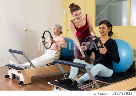 Hispanic woman exercising with flex ring and fitball on reformer with young instructor 112788932