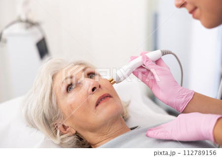 Young female dermatologist performing cosmetology procedure for old woman, RF lifting and vacuum massage for facial skin therapy in clinic 112789156