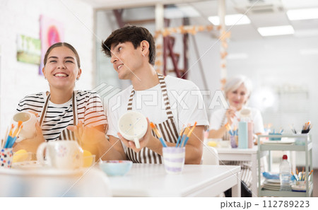 Cheerful young couple decorating ceramic pottery at table in workshop Cheerful young couple decorating ceramic pottery at table in workshop 112789223