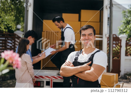 Portrait of a smiling mover unloading boxes into a new home from a truck. These removal company workers guarantee a joyful relocation and efficient service. Moving day concept Portrait of a smiling mover unloading boxes into a new home from a truck. These removal company workers guarantee a joyful relocation and efficient service. Moving day concept 112790312