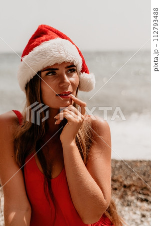 Woman summer travel sea. Happy tourist in red bikini and Santas hat enjoy taking picture outdoors for memories. Woman traveler posing on the beach surrounded by volcanic mountains, sharing travel joy 112793488
