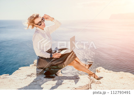 Digital nomad, Business woman working on laptop by the sea. Pretty lady typing on computer by the sea at sunset, makes a business transaction online from a distance. Freelance, remote work on vacation 112793491