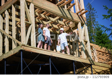 Man architect showcases to couple process of constructing wooden framed house. Youthful pair of investors inspecting their future dwelling in mountains. Concept of contemporary ecological construction Man architect showcases to couple process of constructing wooden framed house. Youthful pair of investors inspecting their future dwelling in mountains. Concept of contemporary ecological construction 112793909