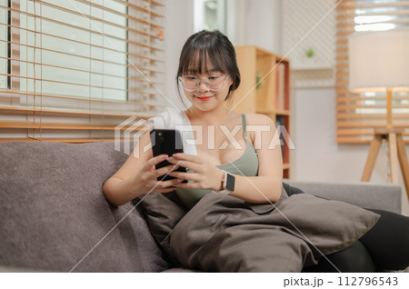A woman is using her phone and relaxing on the sofa in her living room after finishing her workout. 112796543