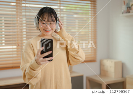 A cute Asian woman is listening to music on her headphones and using her phone in the living room. A cute Asian woman is listening to music on her headphones and using her phone in the living room. 112796567