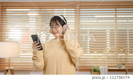 A cute Asian woman is listening to music on her headphones and using her phone in the living room. 112796574